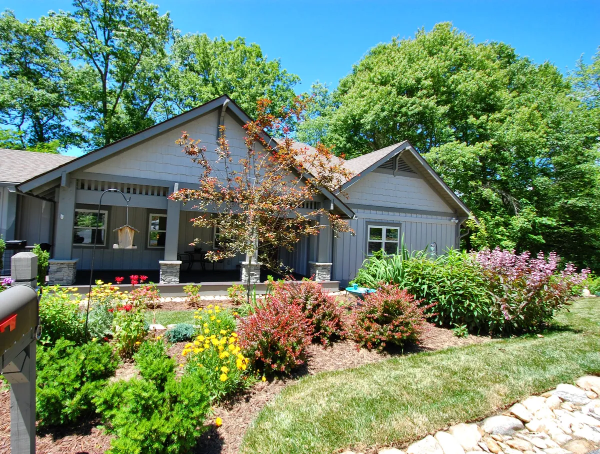 A charming view of the cottage’s front porch framed by lush green trees and vibrant flower beds. The inviting porch offers a tranquil space for morning coffee or winding down after a productive day. The surrounding nature is visible, suggesting a peaceful connection to the outdoors and proximity to walking trails and the Highlands neighborhood.
