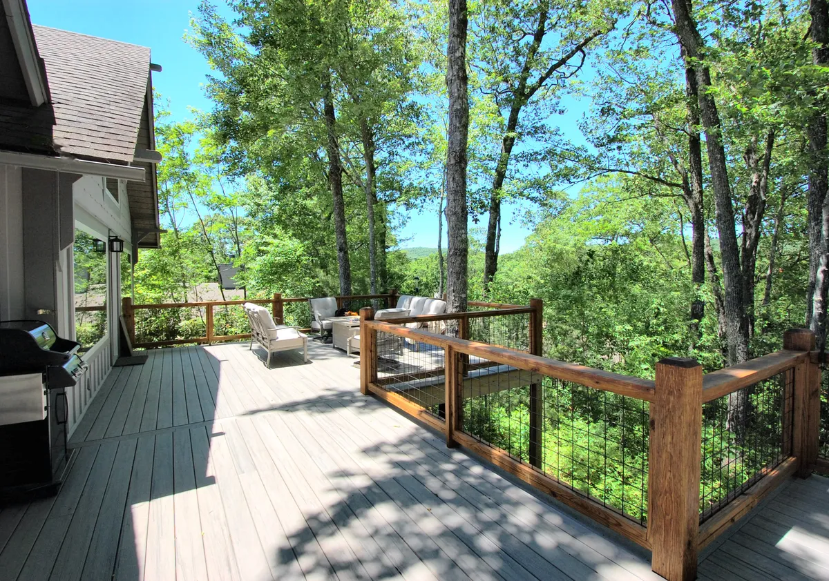 A wide, wooden deck surrounded by lush trees, featuring a spacious outdoor seating area and a grill. The seating area is arranged for both relaxation and social gatherings, and the deck overlooks natural scenery, perfect for enjoying meals or coffee outdoors.