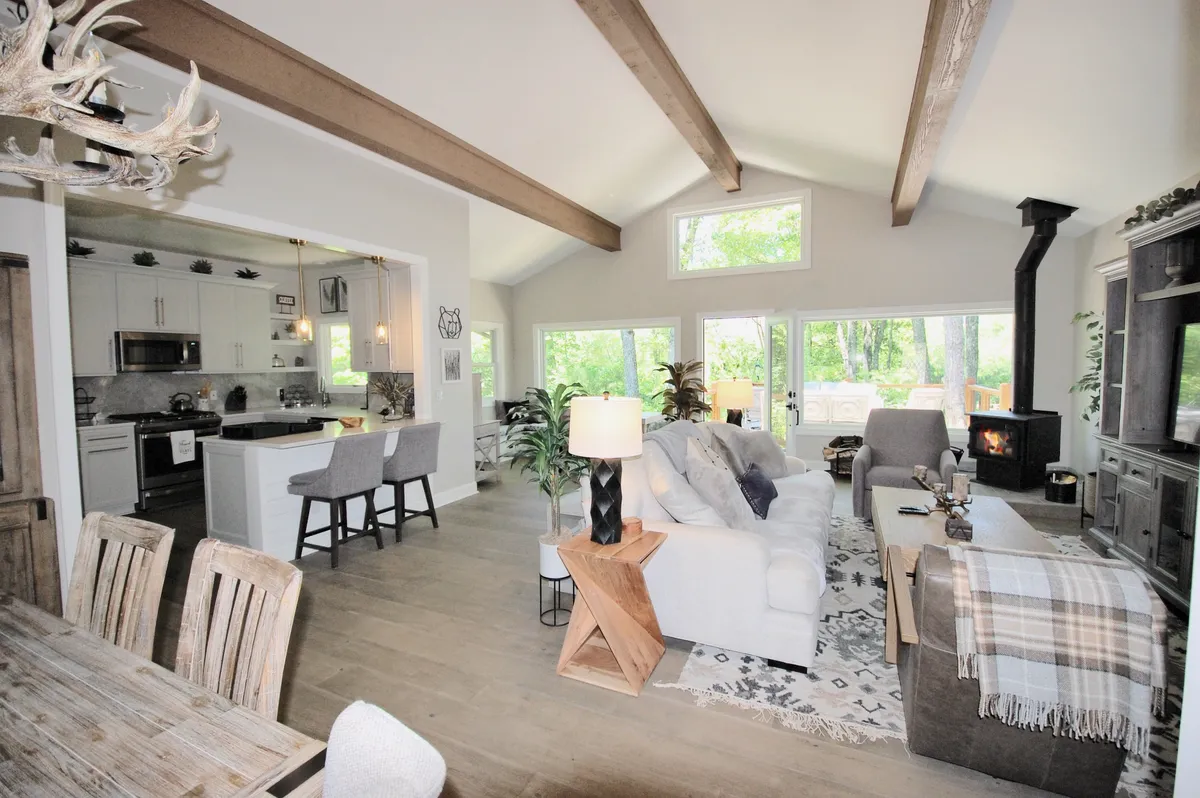 This image presents a spacious, open-plan interior where the living area flows into the dining space. Large windows line the wall, welcoming abundant natural light and offering lush greenery views outside. Modern, comfortable seating surrounds a low coffee table, with a fireplace adding warmth and coziness. A central dining table is positioned conveniently within the same bright, connected space.
