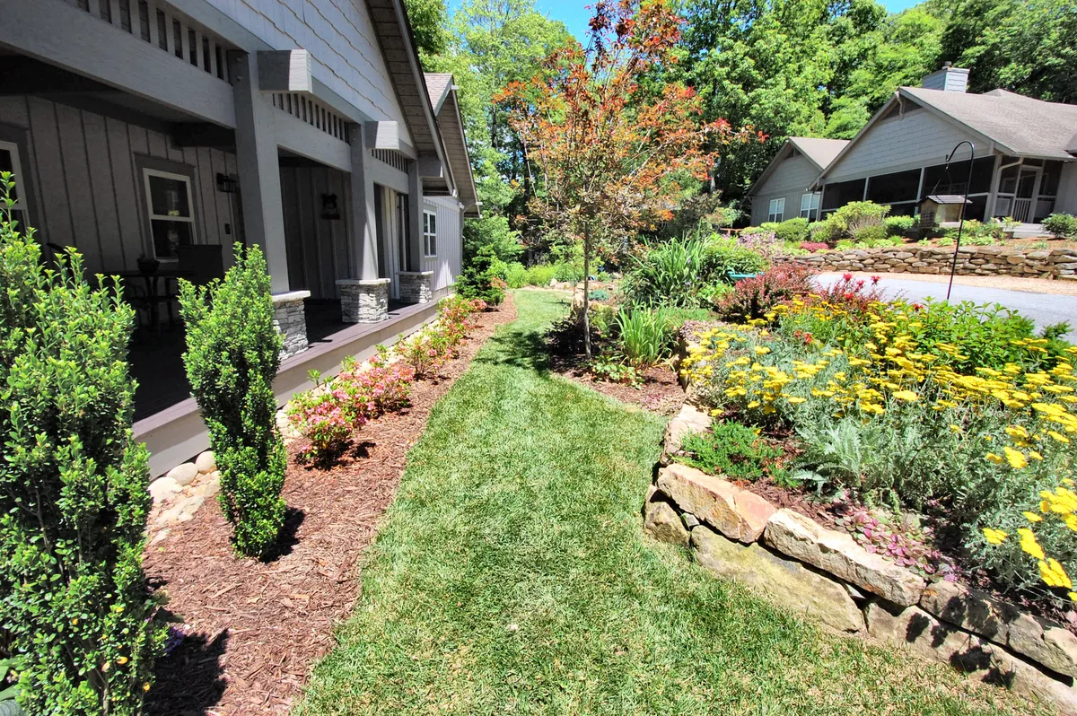 A tranquil garden setting with lush greenery, bright flower beds, a stone pathway, and a tidy lawn creates a serene outdoor environment. The porch in the distance invites morning coffee or sunset relaxation.