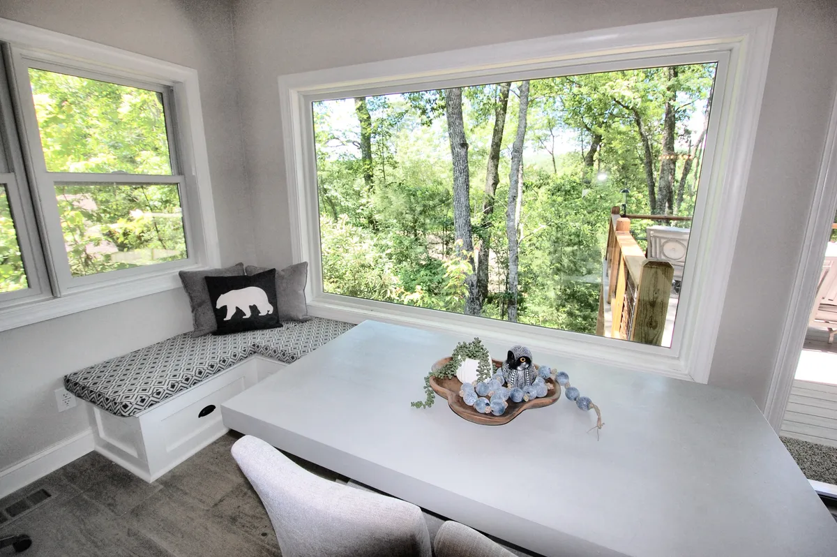 A cozy window seat with a decorative table arrangement sits beside a large window overlooking trees, creating a peaceful nook perfect for casual dining, morning coffee, or quiet reflection. The space feels relaxed and intimate, ideal for cherished moments.